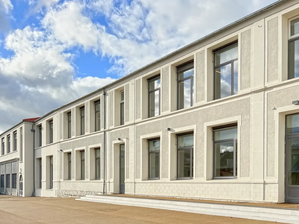 Exterior view of the Groupe Scolaire in Paris, part of a campus of three schools, featuring a two-story cream-colored facade with large wooden-framed windows and doors designed for educational spaces, incorporating sun shading and security protection.